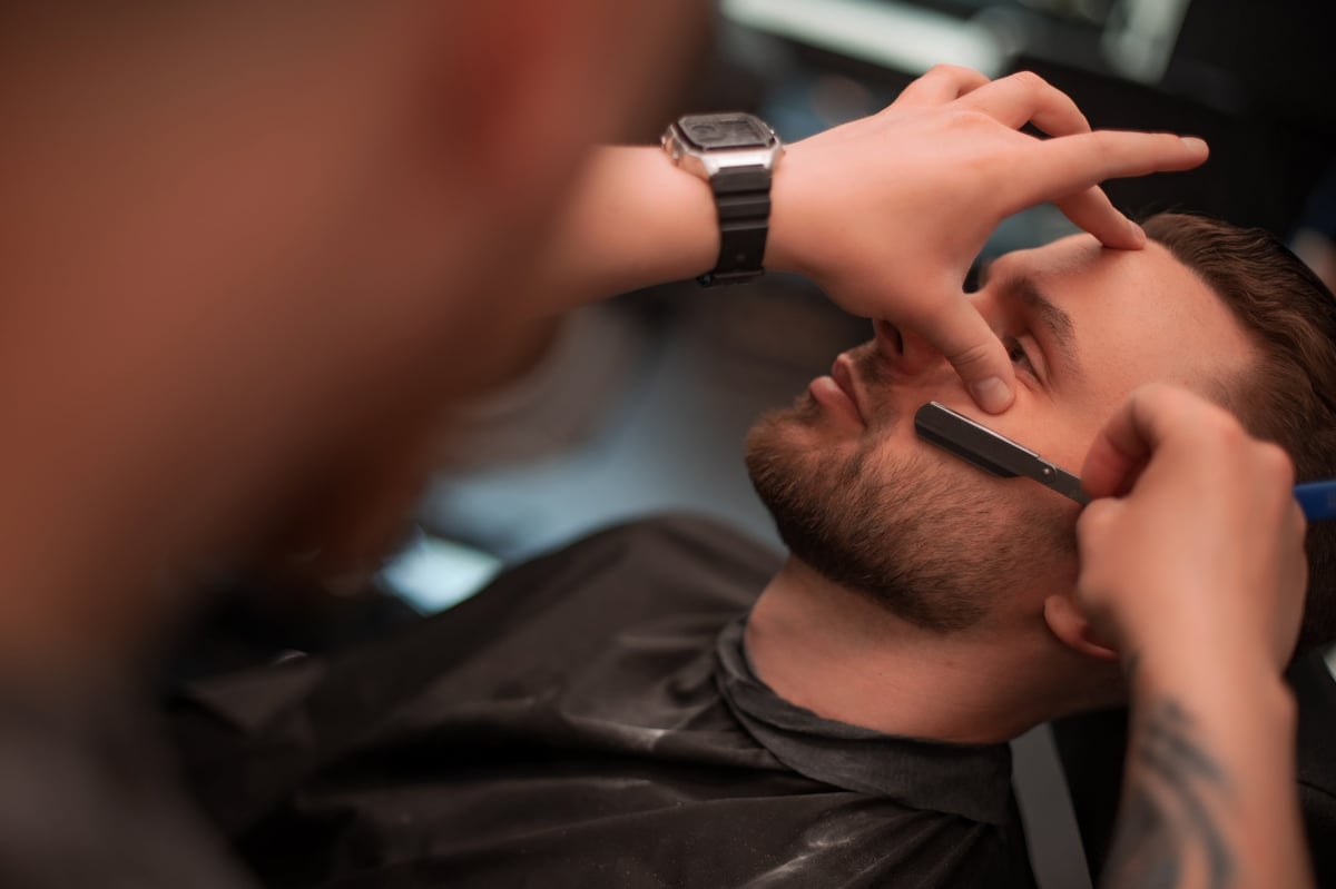 Bearded man reclines in a barber chair receiving a straight-razor shave from a professional barber, showcasing hands, razor, and grooming tools in a modern barbershop setting.